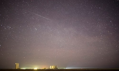 A starry night sky with a faint meteor streaking above a distant landscape illuminated by soft lights.