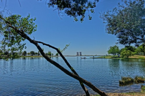 A serene lake scene framed by trees, with cooling towers visible in the distance under a clear blue sky.
