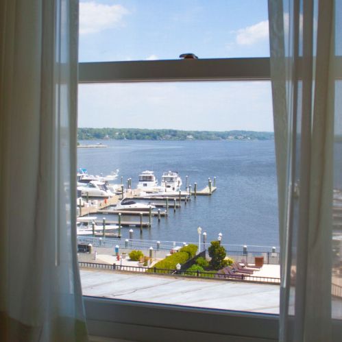 View of a calm waterfront with boats docked, seen through a window with sheer curtains.