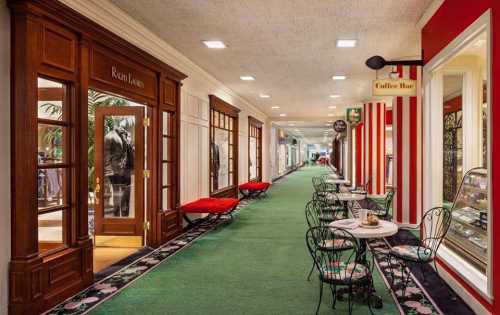 A hallway with green carpet, lined with shops and small tables, featuring a coffee bar and decorative storefronts.