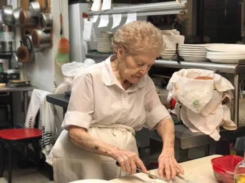 An elderly woman in a white shirt prepares food in a busy kitchen, focused on chopping ingredients.