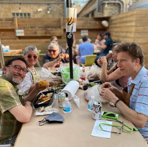 A group of people enjoying a meal outdoors at a table, with food containers and drinks in a casual setting.