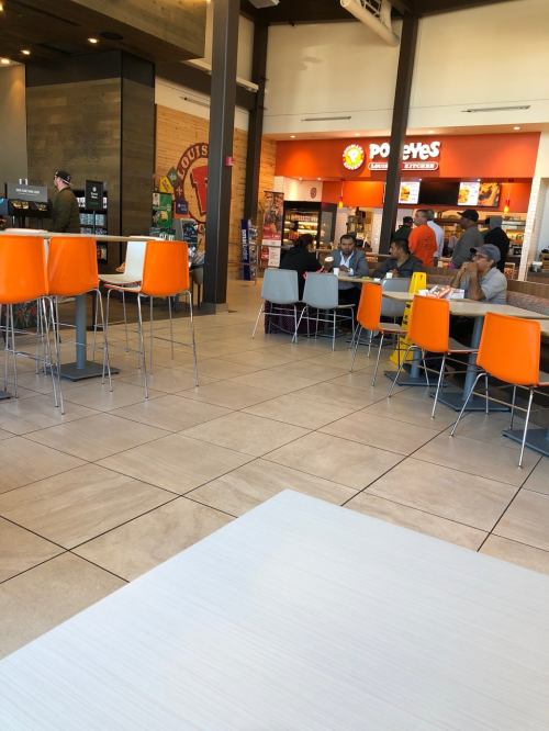 A casual dining area with orange and gray chairs, featuring a Popeyes restaurant in the background and several patrons seated.