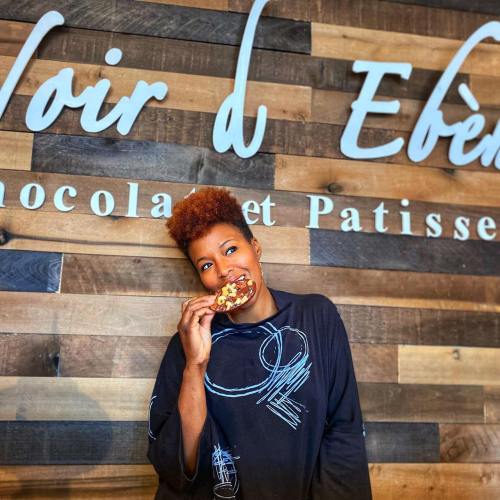 A woman with short, curly hair smiles while holding a cookie in front of a wooden wall with "Noir d'Ébène" signage.