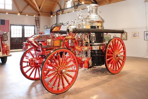 A vintage fire engine with bright red wheels and shiny metal details, displayed in a spacious museum setting.