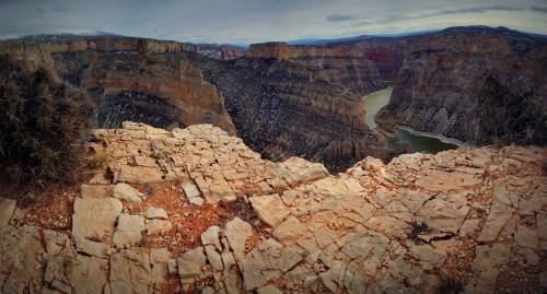 A panoramic view of a canyon with steep rock formations and a winding river below, under a cloudy sky.