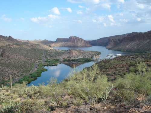 A scenic view of a lake surrounded by mountains and desert vegetation under a blue sky with scattered clouds.