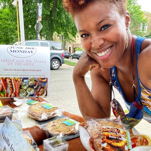 A smiling woman at a food stand with baked goods, promoting a grilling event featuring vegan and vegetarian options.