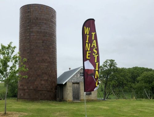 A rustic building with a tall cylindrical structure nearby, featuring a flag that reads "Wine Tasting" in bright colors.
