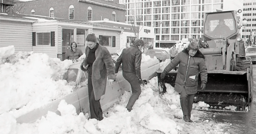 People clearing snow from cars with a bulldozer in a snowy urban setting.