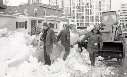 People clearing snow from cars with a bulldozer in a city street during winter. Buildings in the background.