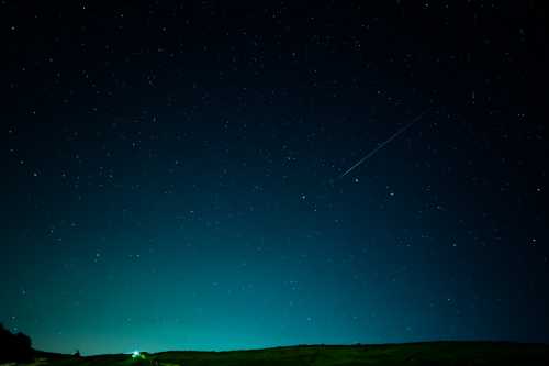 A starry night sky with a faint meteor streaking across, above a dark landscape.