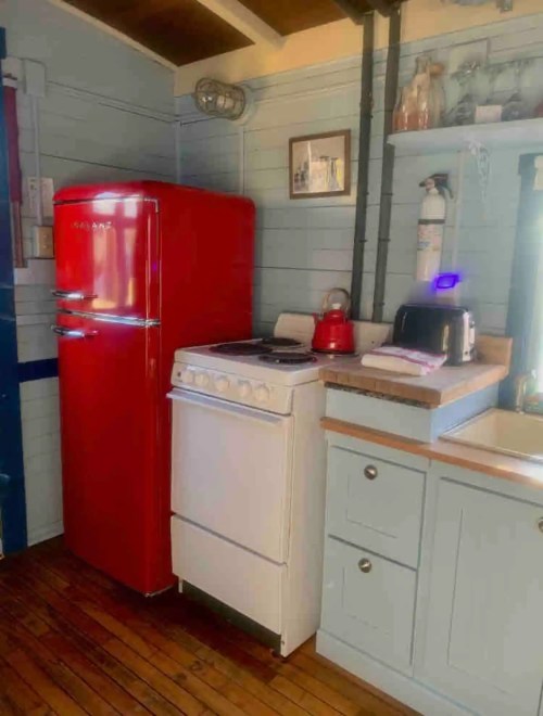 A cozy kitchen featuring a red retro fridge, white stove, kettle, toaster, and light blue cabinets on wooden flooring.