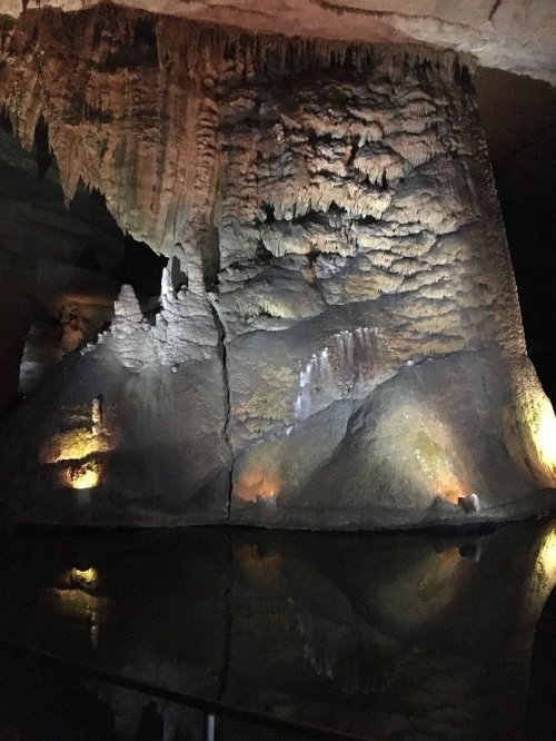 Stalactite-covered cave wall illuminated by lights, reflecting in still water below.