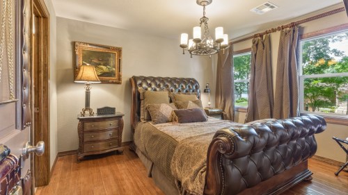 Cozy bedroom featuring a tufted bed, elegant chandelier, and warm decor with natural light from windows.