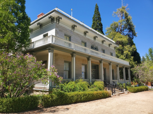 A historic two-story stone house with a balcony, surrounded by greenery and trees under a clear blue sky.