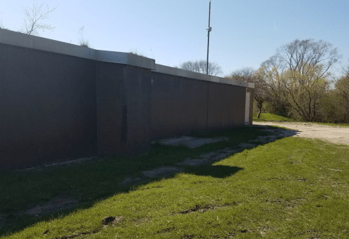 A dark building wall with a grassy area in front, under a clear blue sky. Trees are visible in the background.