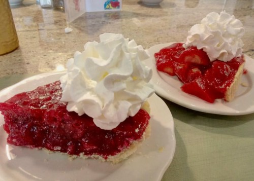 Two slices of cherry pie topped with whipped cream, served on white plates. One slice is whole, the other is partially eaten.