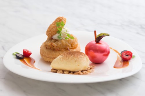 A gourmet dessert plate featuring a pastry, ice cream, a shiny red apple, and garnishes of berries and sauce.