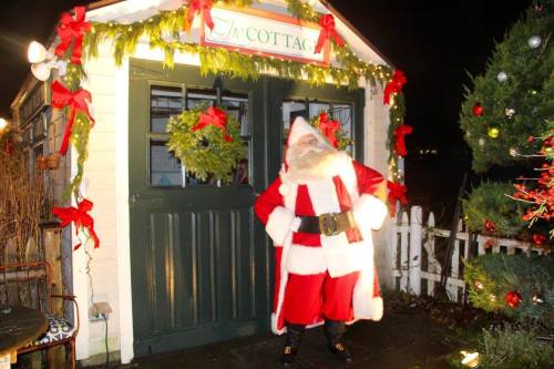 A cheerful Santa Claus stands in front of a decorated cottage, surrounded by festive greenery and holiday lights.
