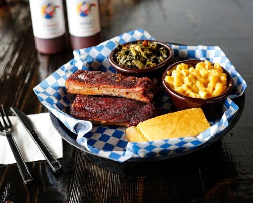 A plate with BBQ ribs, collard greens, macaroni and cheese, and cornbread, served in a blue checkered basket.