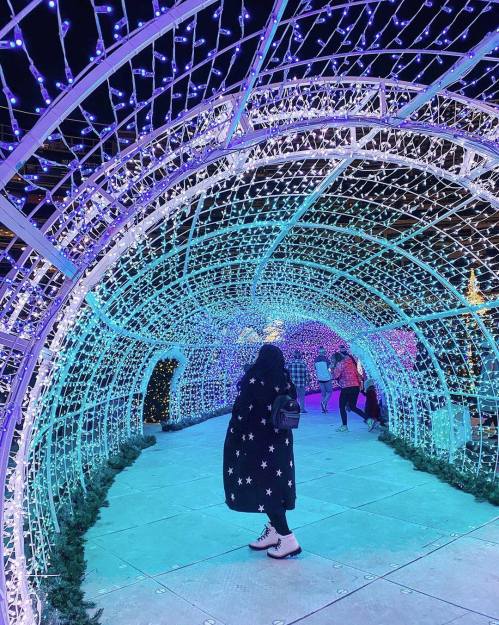 A person in a starry coat walks through a colorful, illuminated tunnel of lights during the night.