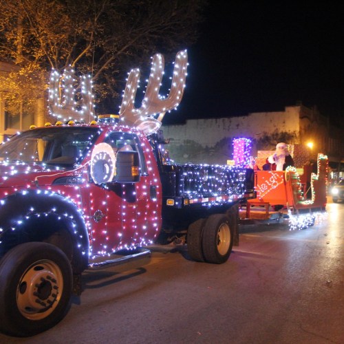 A festive truck adorned with lights and reindeer antlers, pulling a decorated sleigh with Santa.