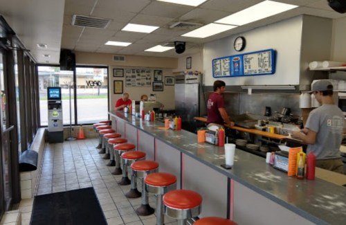 A diner interior with a long counter, red stools, and staff preparing food. Walls adorned with photos and a clock visible.