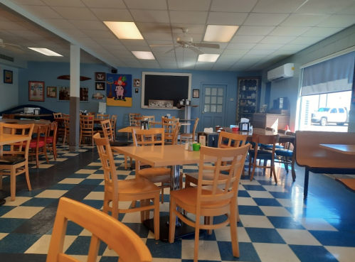 A cozy diner interior with checkered floors, wooden tables, and chairs, featuring blue walls and a chalkboard.