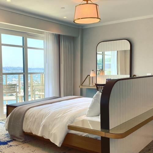 Bright, modern bedroom with a large window, a bed with white linens, and a stylish mirror reflecting a scenic view.