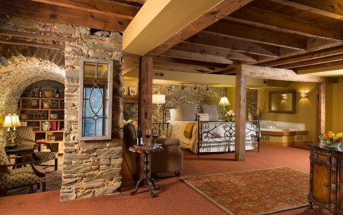 Cozy bedroom with stone walls, wooden beams, and a sitting area, featuring a large bed and a jacuzzi tub.
