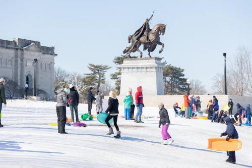 People sledding on a snowy hill near a statue, with children and adults enjoying winter activities in bright clothing.