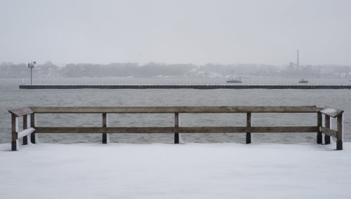 A snowy pier overlooks a calm, gray waterway with distant land and structures shrouded in mist.