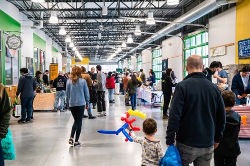 A bustling indoor market with people browsing stalls, colorful decorations, and a child holding a balloon animal.