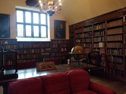 A cozy library with tall bookshelves, a globe, a wooden table, and a red couch under a chandelier.