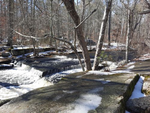 A serene winter scene featuring a flowing river, snow-covered rocks, and bare trees in a wooded area.