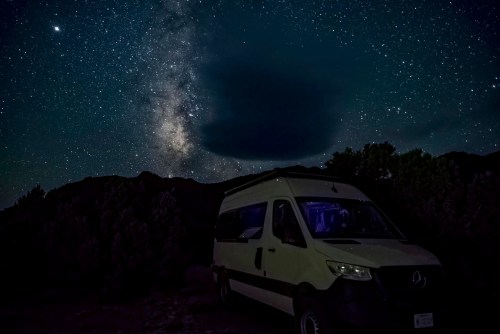 A camper van parked under a starry night sky, with the Milky Way visible and clouds partially obscuring the stars.