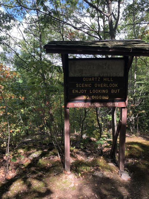 Sign at Quartz Hill Scenic Overlook in Nicolet National Forest, stating "Enjoy looking but no digging." Surrounded by trees.