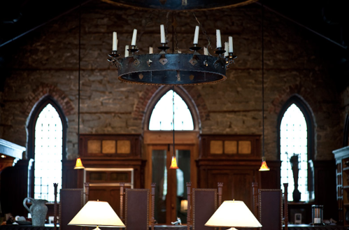 A rustic interior featuring a stone wall, arched windows, and a large chandelier hanging above wooden furniture.