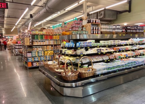 A grocery store aisle filled with shelves of various food products and baskets, featuring a bright, organized layout.