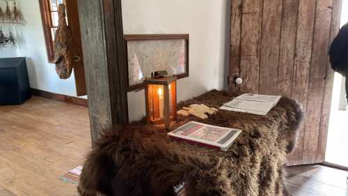 A cozy interior with a wooden table covered in a fur throw, a lantern, and open books near a rustic door.