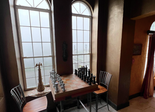 A chess table with black and white pieces, set by large windows with a foggy view outside.