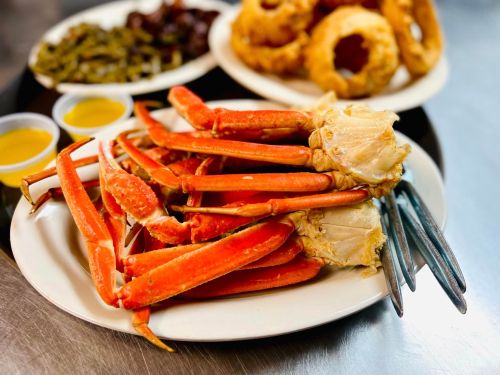 A plate of steamed crab legs with dipping sauces, accompanied by onion rings and sides in the background.
