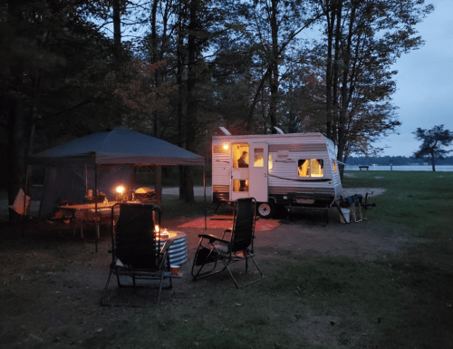 A cozy campsite at dusk featuring a camper, a tent, and chairs around a small fire. Trees surround the area.