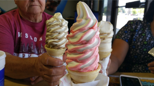 An elderly person holds a soft-serve ice cream cone, surrounded by others with various flavors in a casual setting.