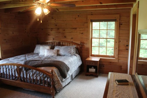Cozy wooden bedroom with a large bed, nightstand, and a window overlooking greenery. Ceiling fan above.