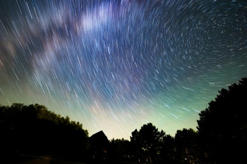 A starry night sky with swirling stars and the Milky Way, framed by silhouettes of trees and a building.