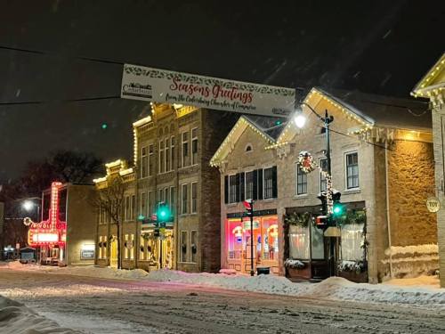 Snowy street scene at night with festive lights on buildings and a "Seasons Greetings" banner overhead.