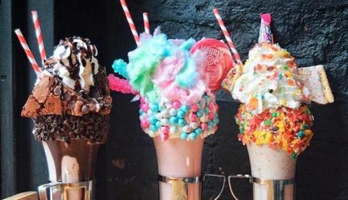 Three colorful milkshakes topped with whipped cream, candy, and sprinkles, served in tall glasses with striped straws.