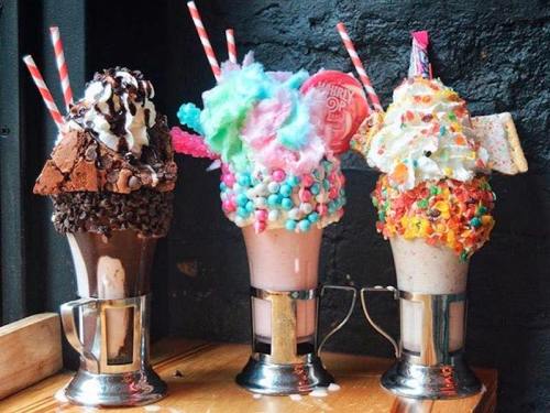 Three extravagant milkshakes topped with colorful candies, whipped cream, and unique garnishes, served in metal glasses.
